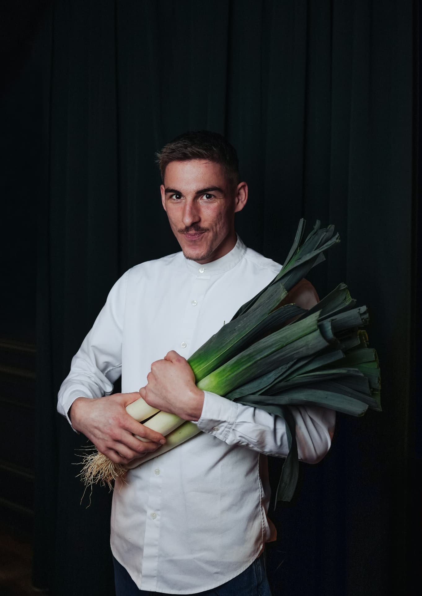 Chef Charlie Anne holding a bundle of fresh leeks against a dark green curtain